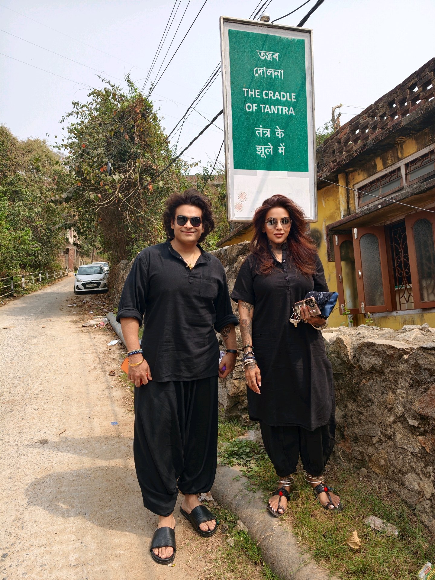 Meenakshii and Punit standing in Kamakhya, Assam, beneath a green road-sign that reads The Cradle of Tantra in Assamese, Hindi and English